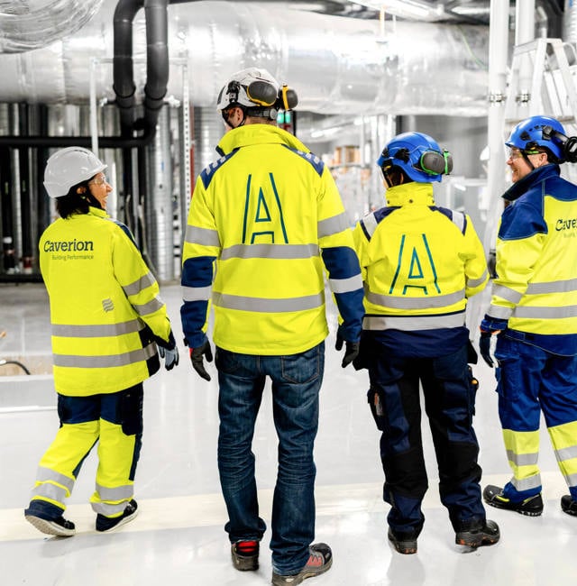 Technicians pistured from behind in a factory looking environment, wearing full safety gear